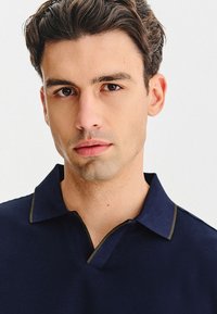 Granatowa koszulka polo z teksturowanej tkaniny, z klasycznym kołnierzem i kontrastującym złotym przeszyciem wzdłuż kołnierzyka i listwy guzikowej.