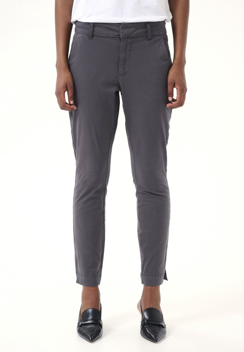 Personne portant un pantalon gris slim et des chaussures noires à bout pointu sans lacets, debout devant un fond blanc uni.