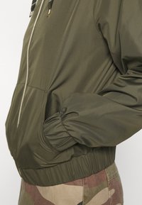Giacca bomber verde oliva con chiusura a zip, polsini elastici e tasche laterali. Realizzata in tessuto morbido e leggero con una vestibilità comoda.