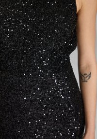 Mustaharjattua mustaa strassipukua, jossa on tiukka muotoilu, esittelee kiiltävän pinnan ja tekstuurisen kuosin. Oikeassa kädessä on näkyvissä tatuointi.