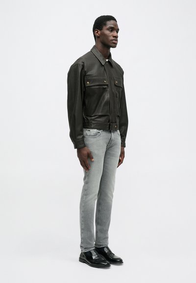 Σκούρο πράσινο δερμάτινο μπουφάν με κουμπωτές στήθους τσέπες, συνδυασμένο με ανοιχτό γκρι τζιν και μαύρα κοστούμια παπούτσια. Το μοντέλο στέκεται μπροστά σε λευκό φόντο.