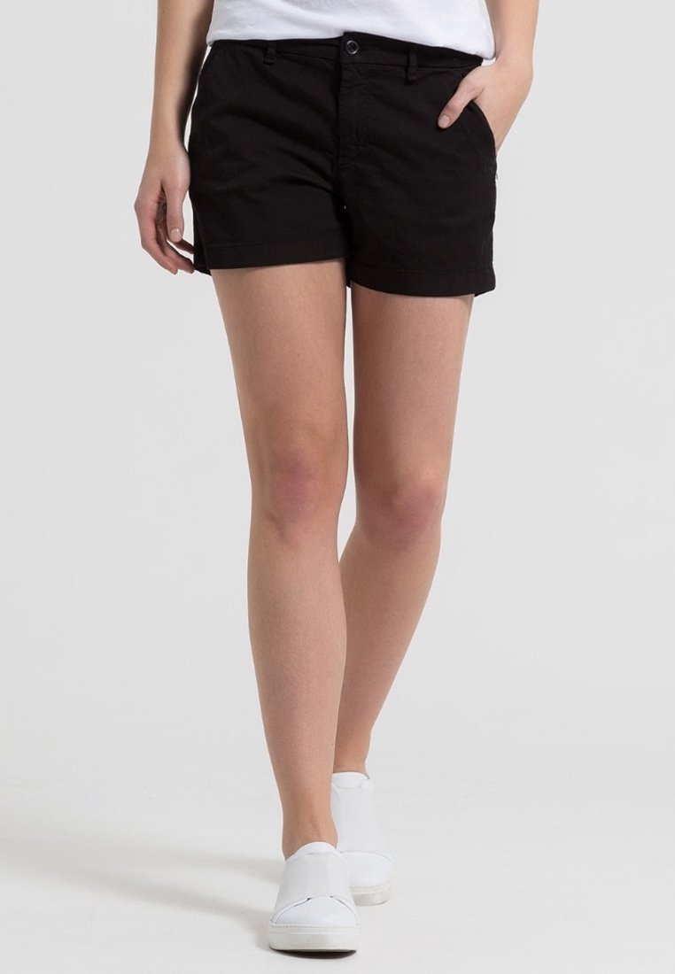 Personne portant un short noir, des chaussures blanches slip-on et une chemise blanche, une main dans la poche, debout devant un fond uni.