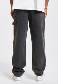 Pantalones cargo de mezclilla negra con pierna recta, bolsillo lateral y dobladillos ajustados, combinados con zapatillas deportivas blancas, mostrados desde la parte posterior.