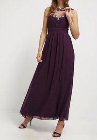 Kvinna iklädd en golvlång, ärmlös mörklila klänning med juvelbesatt halsringning och genomskinlig övre del av bröstet, tillsammans med svarta högklackade sandaler.