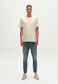 Beige, rövidujjú póló, struktúrált anyagból, kék farmernadrággal és világosszürke, texturált felületű sportcipővel párosítva.