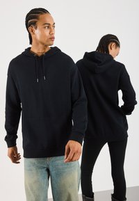 Svart hettegenser med kengurulomme, har snøring og ribbede mansjetter. Modellen har på seg lys blå jeans og mørke bukser.
