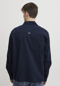 Chemise à manches longues bleu marine avec un col, des poignets boutonnés et une petite étiquette au dos. Fabriquée dans un tissu lisse et durable.