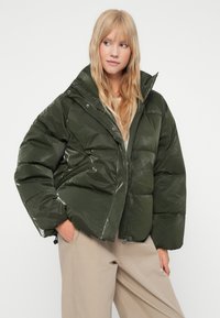 Puffet grøn jakke lavet af skinnende materiale, med høj krave, lynlåslukning og sidelommer. Designet er oversized med en quiltet tekstur.