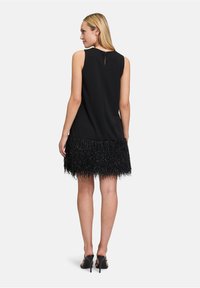 Czarna sukienka bez rękawów z teksturowanym, frędzlonym dołem. Posiada dekolt w kształcie łezki z tyłu oraz gładki materiał, zestawiona z czarnymi sandałami na obcasie.