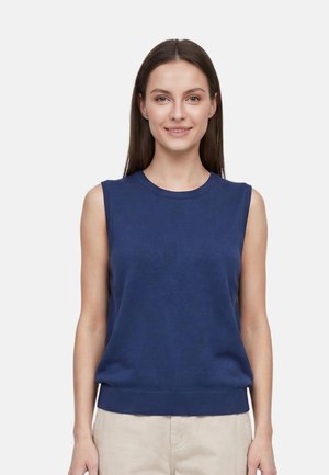 Giovane donna con capelli castani lisci che indossa una canotta blu navy senza maniche e pantaloni beige, sorridente e rivolta verso avanti su uno sfondo bianco.