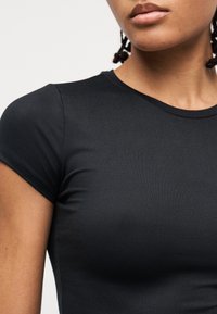 Κοντινό πλάνο ενός ατόμου που φοράει απλό, μαύρο, εφαρμοστό κοντομάνικο τοπ, δείχνοντας το πάνω μέρος του στήθους, τον ώμο και τα χεί λη.