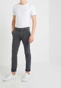 Man i en enkel vit t-shirt med kort ärm, mörkgrå avslutade byxor och vita sneakers, står med händerna i fickorna.