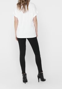 Donna con capelli lunghi che indossa una maglietta bianca a maniche corte, pantaloni skinny neri e stivaletti neri con tacco alto, di spalle.