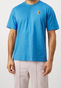 Man wearing a bright blue short-sleeve T-shirt with a small multicolored logo on the chest and light pink pants with seam details.