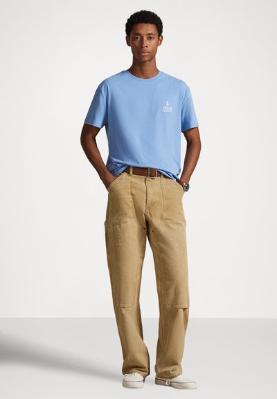 Γαλάζιο βαμβακερό t-shirt με μικρό λευκό λογότυπο στο στήθος, συνδυασμένο με μπεζ παντελόνι εργασίας με τσέπες και ελαφρύ φθαρμένο εφέ.