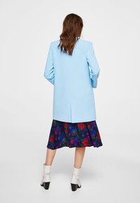 Donna in piedi con le spalle rivolte, indossa un soprabito blu chiaro, una gonna midi floreale e stivali bianchi alla caviglia su uno sfondo semplice.