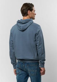 Šedá bavlnená mikina s voľným strihom, s kapucňou na šnúrky a rebrovanými manžetami, v kombinácii s modrými denimovými džínsy.