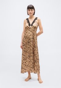 Γυναίκα στέκεται ξυπόλητη φορώντας ένα μαν sleeveless αμάνικο μεταξωτό φόρεμα με λεοπάρ σχέδιο και μαύρη δαντέλα σε απλό φωτεινό φόντο.