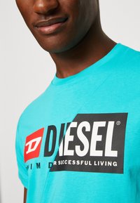 Turquoise cotton T-shirt featuring a large black and white "DIESEL" logo with a red accent, crew neckline, and smooth texture.