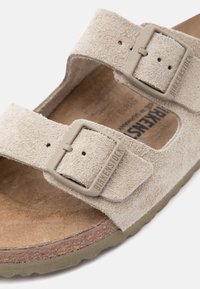 Πέδιλο Birkenstock από μπεζ σουέντ με δύο ρυθμιζόμενα λουράκια με αγκράφες και διαμορφωμένο τακούνι από φελλό σε λευκό φόντο.