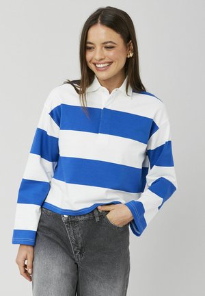 Femme souriante, portant une chemise à manches longues rayée bleu et blanc et un jean gris, debout avec une main dans la poche sur un fond clair.