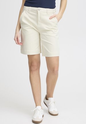 Shorts beige chiaro in cotone, con parte anteriore liscia, due tasche laterali e orli arrotolati, abbinati a sneakers bianche.