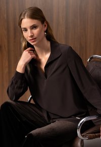 Jeune femme aux longs cheveux portant un chemisier sombre et ample ainsi qu'un pantalon, assise sur une chaise avec un fond en panneaux de bois.