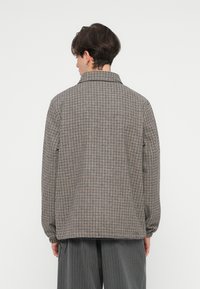 Jachetă în carouri de culoare marron, crem și negru. Prezintă un guler, mâneci lungi și manșete elastice. Materialul pare texturat și ușor supradimensionat.