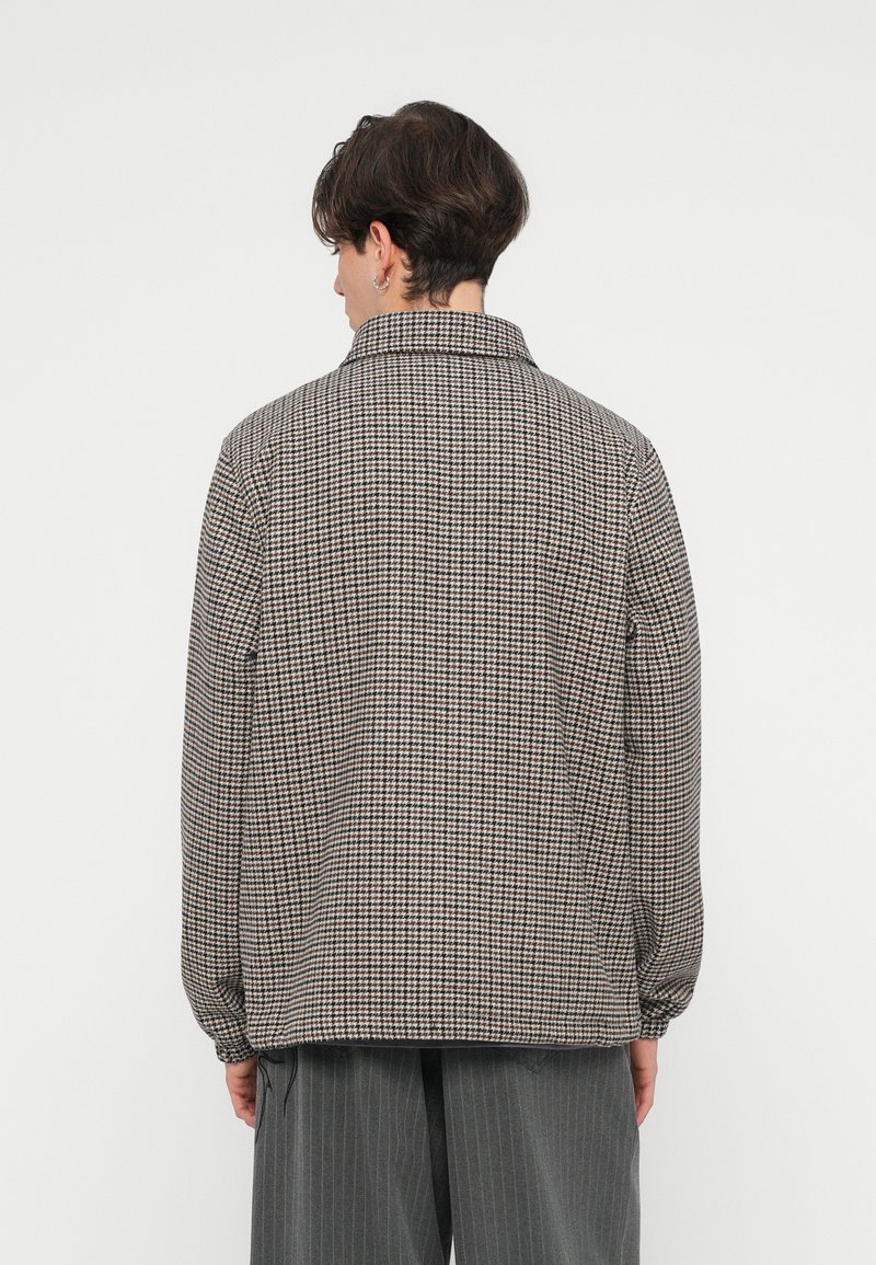 Jachetă în carouri de culoare marron, crem și negru. Prezintă un guler, mâneci lungi și manșete elastice. Materialul pare texturat și ușor supradimensionat.