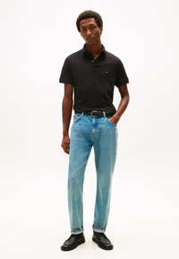 Polo nera, jeans azzurri a gamba dritta con leggero effetto sbiadito, cintura nera testurizzata e scarpe nere. Modello in piedi con le mani nelle tasche.