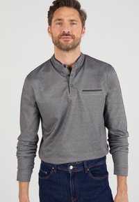 Grijze polo met lange mouwen en een Mandarin-kraag, één borstzak en contrasterende stiksels. G gedragen met blauwe denim jeans.
