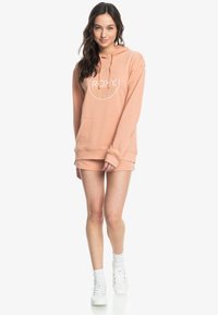 Giovane donna standing in felpa pescato e pantaloni corti abbinati con sneakers bianche, sorridente e rivolta in avanti su uno sfondo bianco.