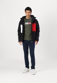 Veste isolée noire et rouge avec fermeture éclair, dotée d'une capuche et d'un design color block. Associée à un t-shirt vert avec logo et un jean foncé.