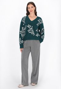 Teal színű kötött pulóver V-nyakkal és ezüst falevél mintázattal, szürke bőszáru nadrággal és világos színű lapos cipőkkel párosítva, fehér háttér előtt.
