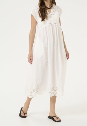 Femme portant une robe midi blanche avec des détails en dentelle ajourée et des tongs noires, debout devant un fond blanc uni.