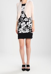 Femme portant une robe noire avec un grand imprimé floral blanc et un cardigan court rose clair, assortie de talons noirs.
