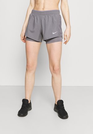 Persoon die grijze dubbellaagse Nike hardloopshorts en zwarte sportschoenen draagt, staand tegen een effen lichte achtergrond.