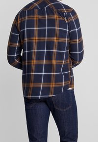 Chemise à carreaux bleu marine avec des lignes orange et blanches, dotée d'un col boutonné et de manches longues, portée avec un jean foncé.