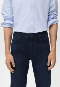 Camisa de manga larga azul claro con las mangas arremangadas, metida dentro de unos vaqueros oscuros con bolsillos frontales y pasadores para el cinturón.
