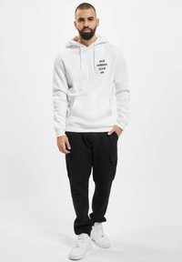 Weißer Kapuzenpullover mit "ANTI CORONA CLUB DEF" in schwarzer Schrift, Kängurutasche und schwarze Cargo-Hosen, kombiniert mit weißen Sneakers.