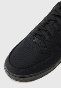 Svarte joggesko med et teksturert stoffoverdel, flate svarte lisser og en gummisole med et mønstret såle. Pyntet med en liten logo.