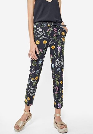 Femme portant un pantalon slim à imprimé floral noir, un haut noir sans manches et des sandales à plateforme beige avec brides à la cheville.