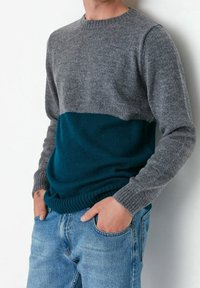 Gebreide trui met een grijs bovenstuk en een donkerblauw/groen onderstuk. Heeft ribgebreide manchetten en een zoom, en een ronde halslijn. Draagt over een blauwe spijkerbroek.