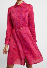 Abito camicia rosa a maniche lunghe con colletto, caratterizzato da un motivo ondulato testurizzato, chiusura con bottoni frontale e cintura abbinata con fibbia dorata.