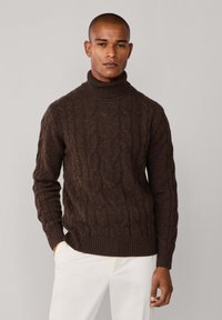 Jersey de tortuga de punto trenzado marrón con puños y dobladillo acanalados, llevado sobre pantalones blancos, mostrando un diseño texturizado.