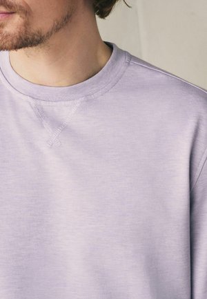 Mann trägt ein hellviolettes Sweatshirt mit Rundhalsausschnitt und sichtbaren Nahtdetails am Kragen vor neutralem Hintergrund.