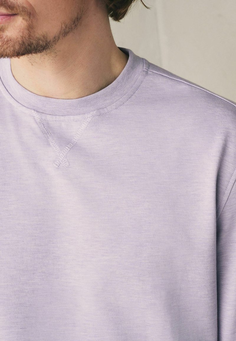 Homme portant un sweat-shirt à col rond violet clair avec un détail de couture visible près du col sur un fond neutre.