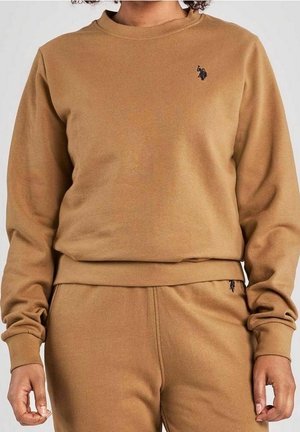 Person iført matchende lysebrun sweatshirt og sweatpants med lille mørkt broderet logo på bryst og hofte.