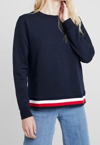 Felpa navy con maniche lunghe, scollo rotondo e orlo con strisce rosse, bianche e navy. Tessuto morbido in misto cotone con vestibilità rilassata.