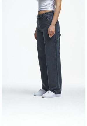 Persoon die zwarte wijde jeans, witte sneakers en een wit cropped topje draagt, staand tegen een effen witte achtergrond.